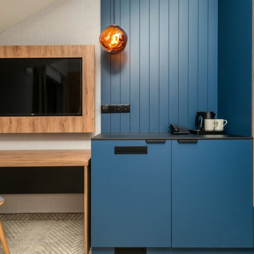 Moderner Hotelbereich mit Holzschreibtisch und Stuhl, blauen Schränken, Wandfernseher und Kaffeemaschine.