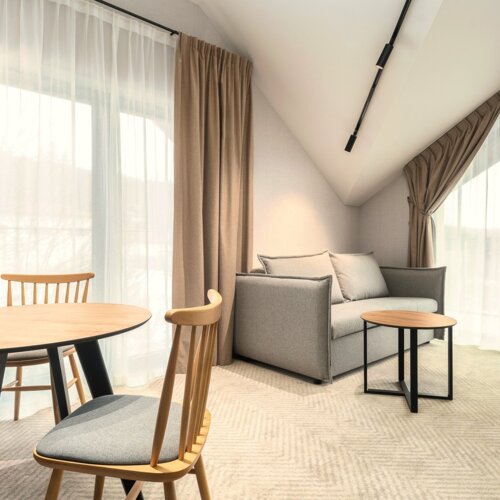 Heller Hotelraum mit schräger Decke, grauem Sofa, rundem Couchtisch, Holztisch und Stühlen vor großen Fenstern mit Vorhängen.