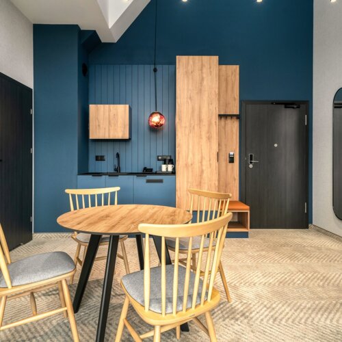 Modernes Hotelzimmer mit Essbereich, Holztisch, vier Stühlen, blauer Akzentwand und kleiner Küchenzeile