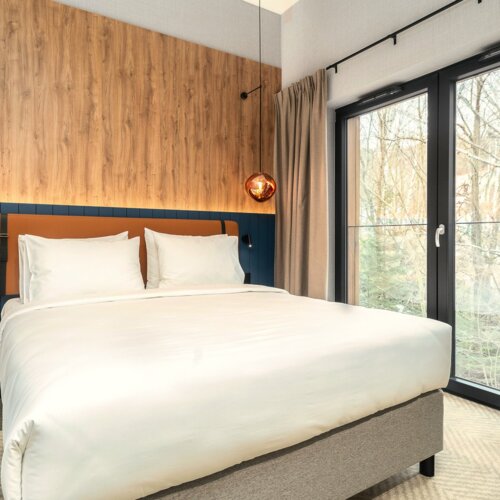 Modernes Hotelzimmer mit großem Bett, Holzverkleidung, hängenden Pendelleuchten und bodentiefem Fenster mit Vorhängen