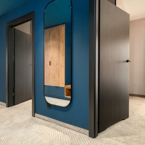 Moderne Hotelsuite mit dunkelblauer Wand, großem Spiegel, Holzstühlen und beigen Vorhängen für komfortablen Aufenthalt.