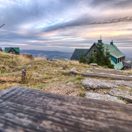 Berghütte mit grünem Dach an Wanderweg mit Panoramablick ins Tal bei Dämmerung und bewölktem Himmel.