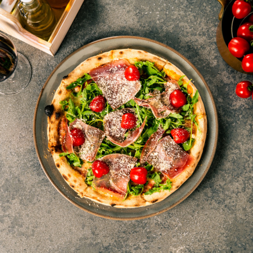 Pizza mit Rucola, Prosciutto und Kirschtomaten auf einem Teller serviert mit einem Glas Rotwein