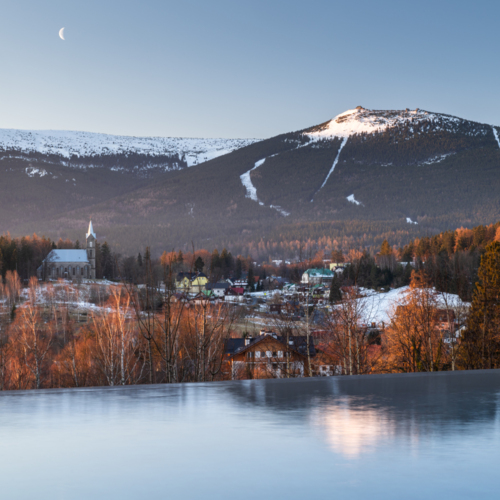 Infinity-Pool mit Blick auf schneebedeckte Berge, malerisches Dorf mit Kirche und herbstlichen Bäumen bei Dämmerung