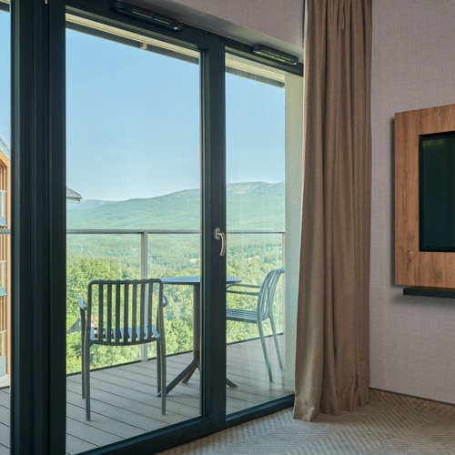 Hotelzimmer mit Balkon, Tisch und Stühlen, Blick auf grüne Berglandschaft und benachbartes Holzgebäude