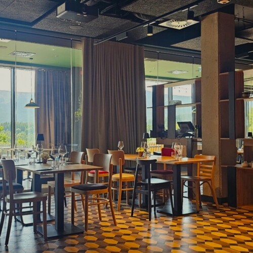 Helles Hotelrestaurant mit Holztischen und Stühlen, großen Fenstern mit Bergblick und Terrasse im Freien.
