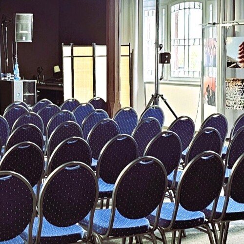 Konferenzraum mit blauen Stühlen in Reihen, Beamer und Fotoausstellung bei den Fenstern