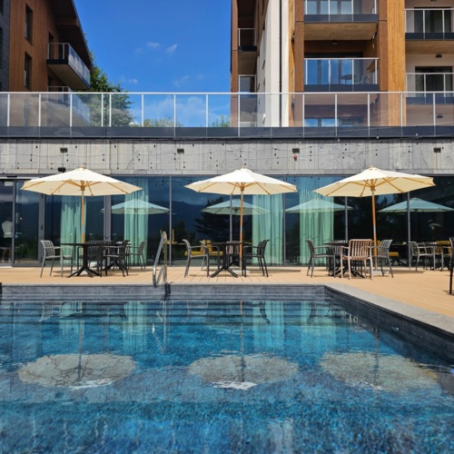Hotelterrasse am Pool mit drei Tischen, Stühlen und Sonnenschirmen vor einem modernen Gebäude mit Holzdetails.