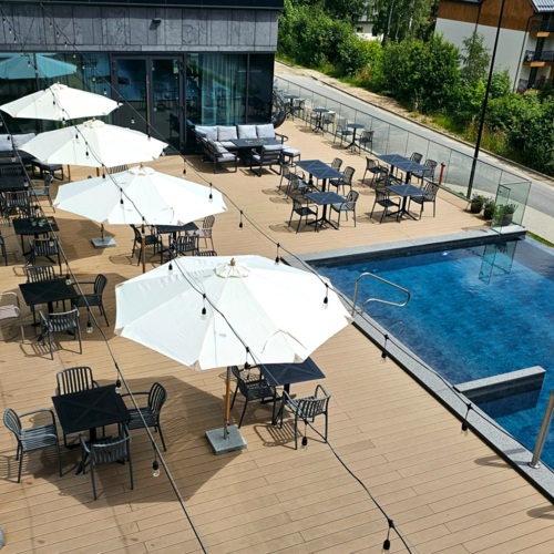 Hotelterrasse mit Außenpool, schwarzen Stühlen und Tischen, beschattet von großen weißen Sonnenschirmen.