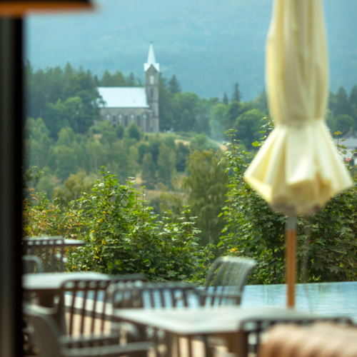 Außenterrasse eines Restaurants mit Metalltischen und Stühlen, mit Blick auf grüne Bäume und eine Kirche auf einem Hügel