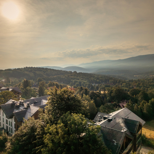 Bergresort mit modernen Häusern zwischen grünen Bäumen und nebligen Hügeln im warmen Sonnenlicht.