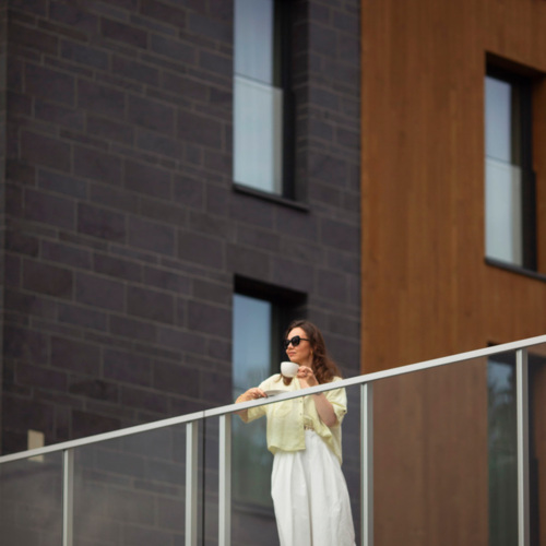 Gast entspannt auf Balkon mit Glasgeländer eines modernen Hotels mit dunkler Stein- und Holzfassade.