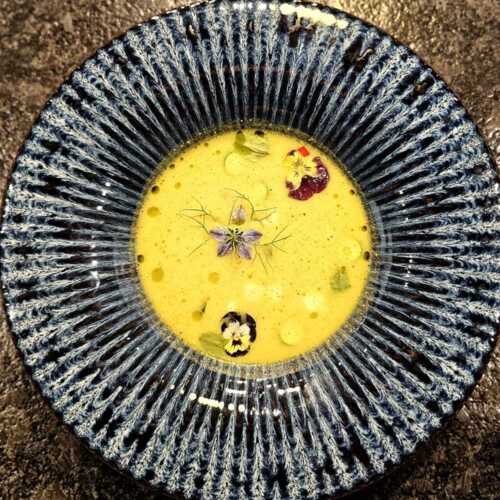 Cremige gelbe Suppe in dekorativer blauer Schale, garniert mit essbaren Blumen und Kräutern als elegante Vorspeise