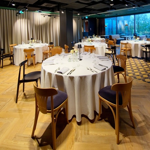 Moderner Speisesaal im Hotel mit runden Tischen, weißen Tischdecken, Holzstühlen und großen Fenstern mit Blick ins Grüne.