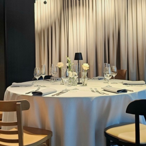 Runder Tisch mit weißem Tischtuch, Weingläsern, weißen Blumen und sanfter Beleuchtung in einem stilvollen Hotelrestaurant.