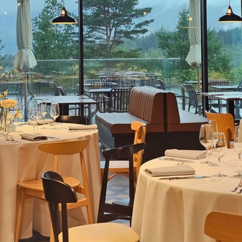 Elegantes Restaurant mit gedeckten Tischen und bodentiefen Fenstern mit Blick auf grüne Bäume.