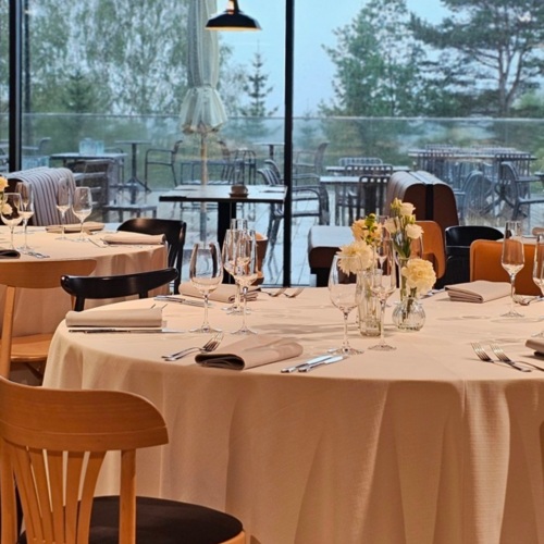 Eleganter Speisesaal mit runden Tischen, weißen Tischdecken, Gläsern und Blick auf die Terrasse mit Bäumen und Gartenmöbeln.