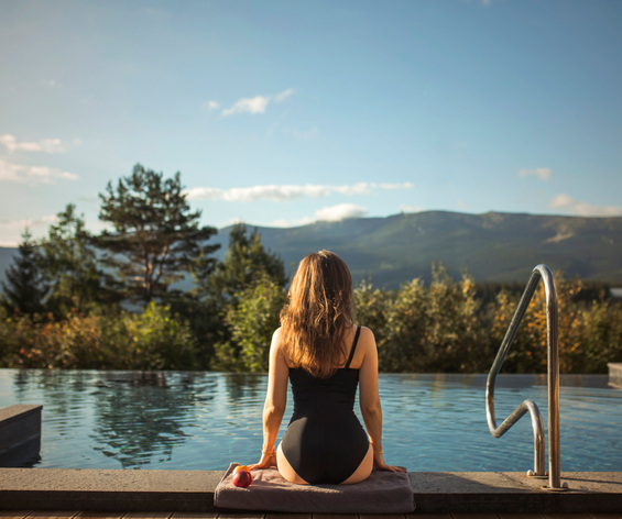 Gast im schwarzen Badeanzug sitzt auf Handtuch am Rand des Infinity-Pools, Blick auf Wälder und Berge, Poolleiter und sanftes Morgenlicht