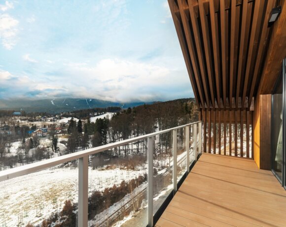 Moderner Hotelbalkon mit Holzfußboden und Glasgeländer, mit Blick auf verschneite Berge und Wald.