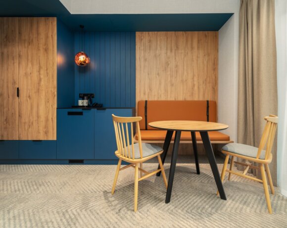 Gemütliche Essecke im Hotelzimmer mit Holzstühlen, orangener Sitzbank, blauer Akzentwand und warmem Hängeleuchter