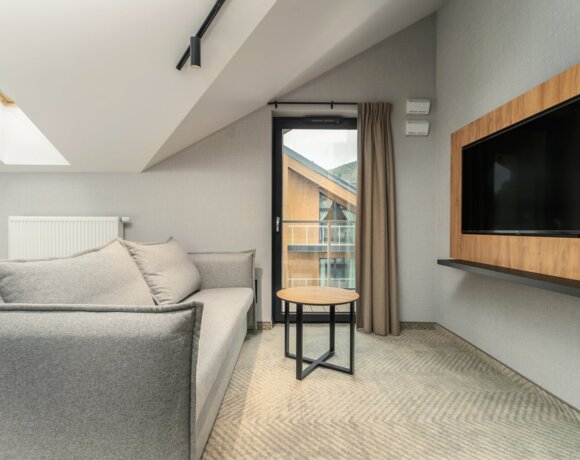 Geräumiges Zimmer mit grauem Sofa, Couchtisch, großem Fernseher und Balkon mit Blick auf Holzgebäude und Berge.