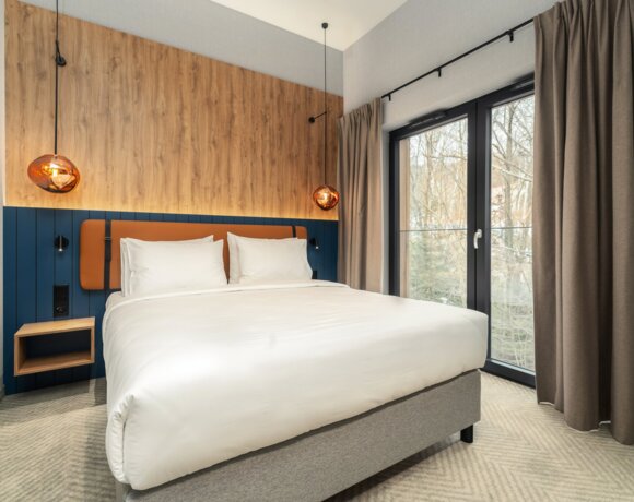 Modernes Hotelzimmer mit großem Bett, dekorativer Holzakzentwand und bodentiefem Fenster mit Blick auf kahle Bäume.