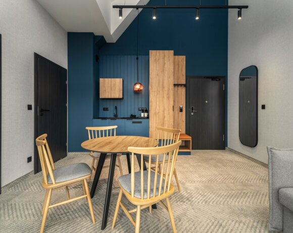 Modernes Hotelzimmer mit blauem Akzentwand, Holz-Esstisch, vier Stühlen und kleiner Küchenzeile