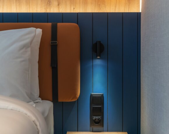 Modern hotel bedroom with blue paneled wall, illuminated wooden accent, and stylish hanging light above a wooden bedside table.