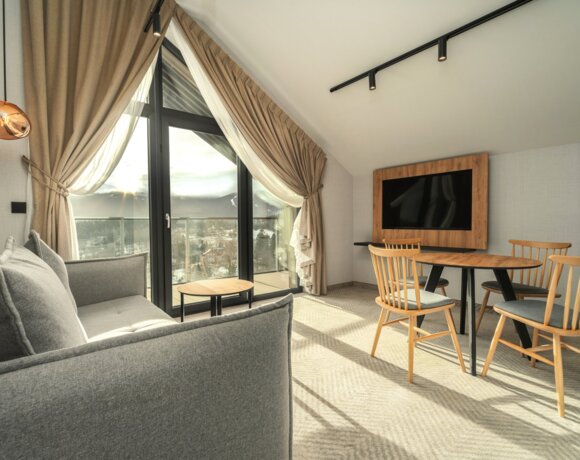 Bright hotel room with large balcony window, gray sofa, wooden dining table, and mountain sunset view in the background.