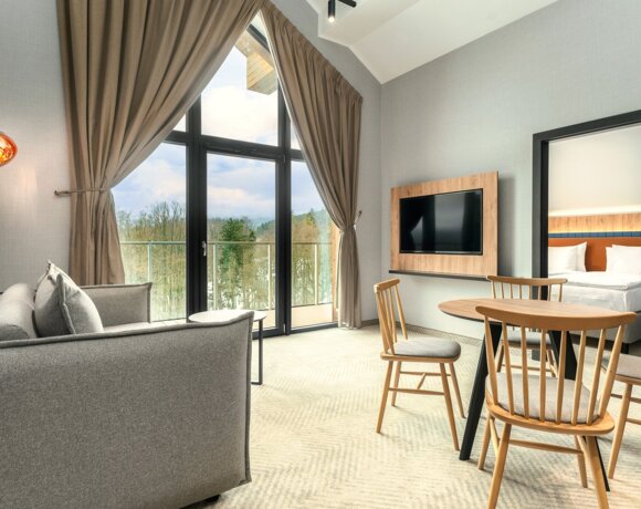 Geräumige Suite mit grauem Sofa, Holzesstisch und Stühlen, Wand-TV und Waldblick durch große Fenster