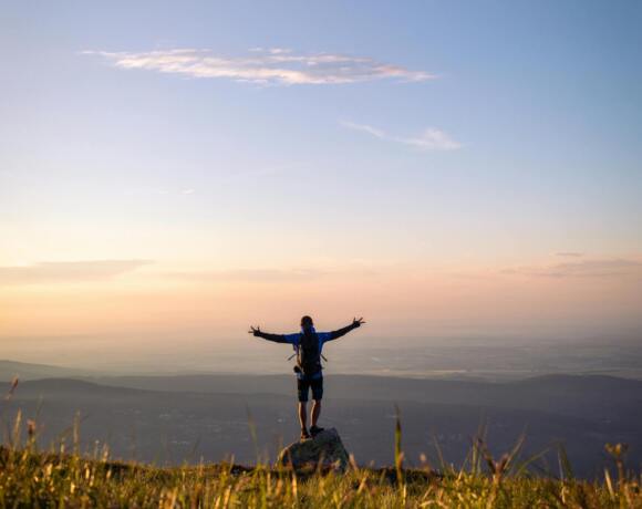Hiker stands on a rock with arms outstretched, overlooking mountain and valley views at sunset.