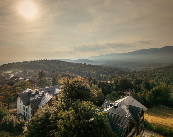 Bergresort mit modernen Häusern zwischen grünen Bäumen und nebligen Hügeln im warmen Sonnenlicht.
