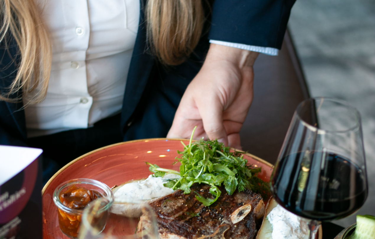 Zdjęcie talerza ze stekiem podawanego przez kelnera na stół, na którym stoją kieliszki wina