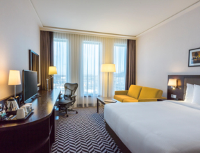 Wnętrze hotelowego pokoju z dwuosobowym łóżkiem, żółtą sofą i miejscem do pracy z obrotowym krzesłem