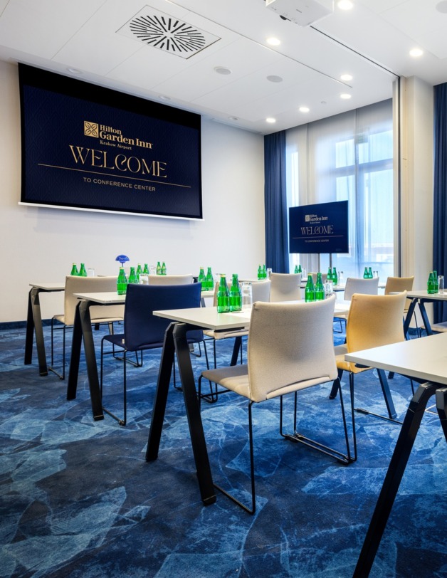 Wnętrze sali szkoleniowej ze stolikami i krzesłami w ustawieniu szkolnym. W tle ekran projekcyjny