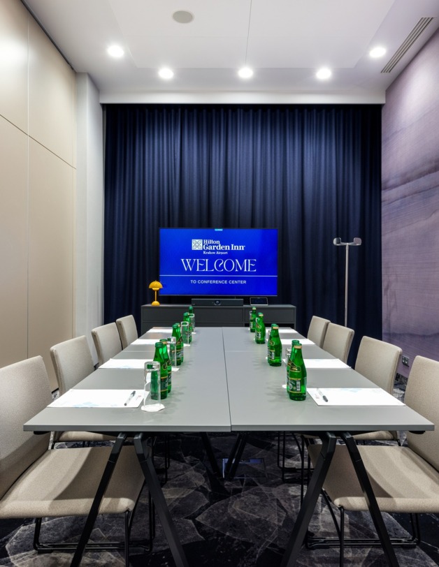 Wnętrze sali ze stołem prezydialnym, na którym stoją butelki z wodą. W tle telewizor