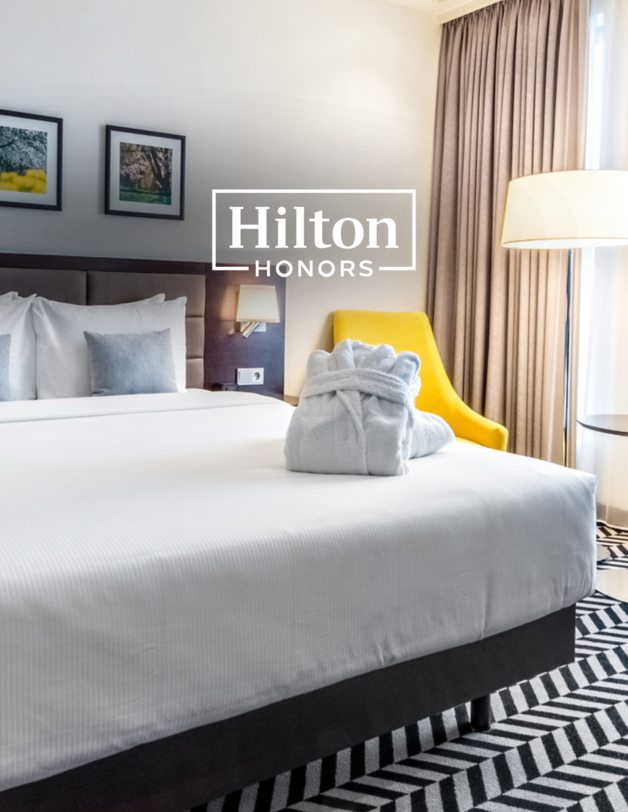 Wnętrze hotelowego pokoju z dużym łóżkiem, na którym leży szlafrok. Logo Hilton Honors