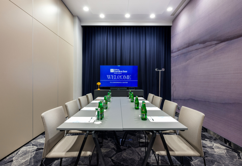 Wnętrze sali ze stołem prezydialnym, na którym stoją butelki z wodą. W tle telewizor