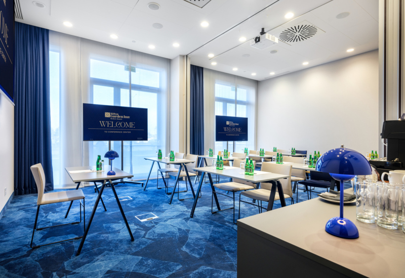 Wnętrze sali szkoleniowej ze stolikami i krzesłami, w tle duże okna