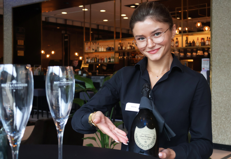 Kelnerka prezentująca butelkę szampana na tle loby baru