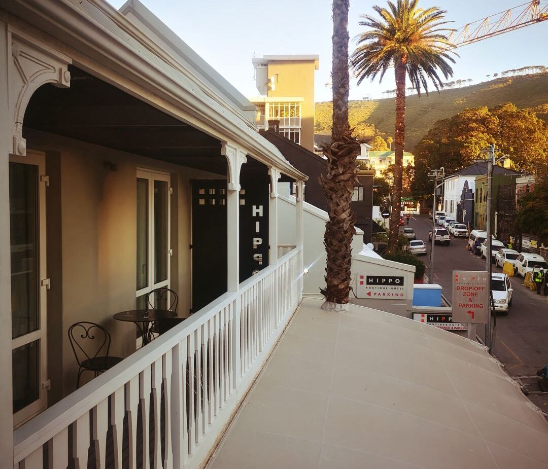 Balcony seating at boutique hotel with white railings, palm trees, and street view with parked cars and hillside backdrop