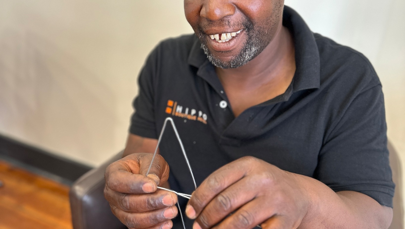 Smiling man in a cap sitting on a leather chair indoors, holding and shaping a thin wire with his hands