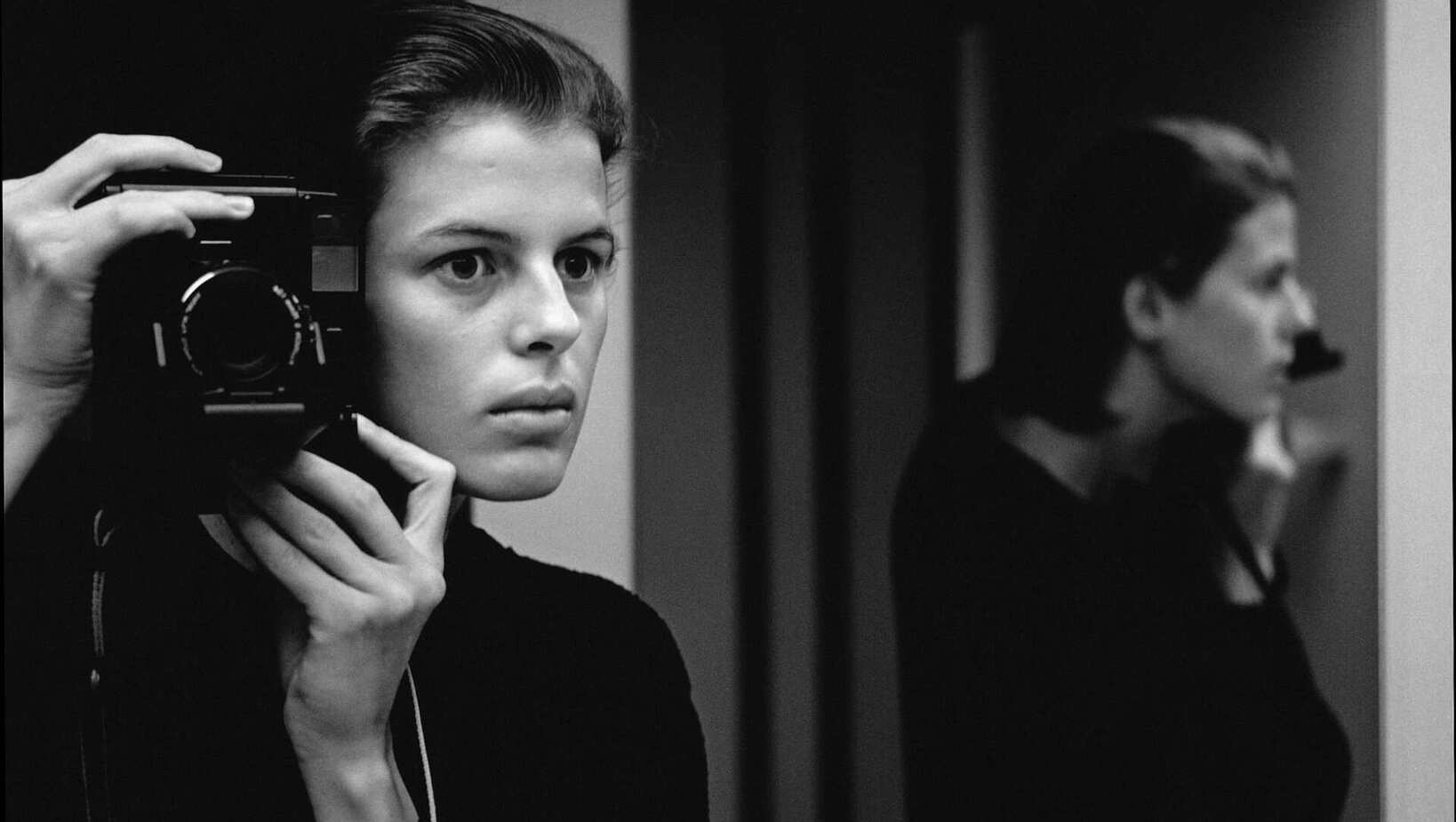 Black and white photo of a woman taking a self-portrait with a camera in a minimalist indoor setting with mirror reflections