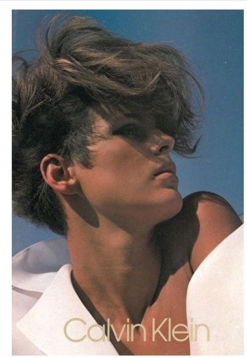 Close-up of a contemplative young person with tousled hair against a clear blue sky, wearing a white shirt collar