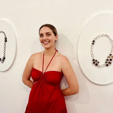 Smiling woman in a red halter dress standing between two minimalist circular art pieces featuring jewelry on a white gallery wall
