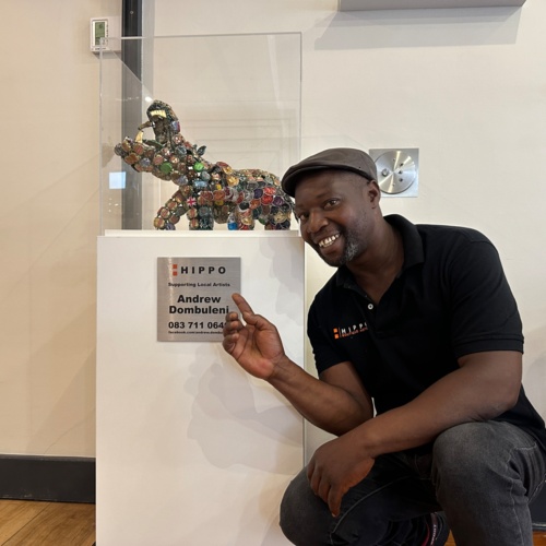 Smiling man in black polo points to hippo sculpture made of colorful bottle caps on white pedestal in hotel lobby