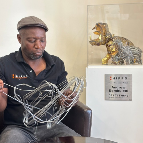 Artist seated indoors crafting a large wire sculpture beside a glass case displaying a metal hippo art piece at Hippo Boutique Hotel