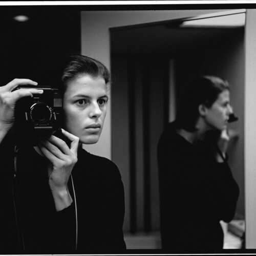 Black and white photo of a woman taking a self-portrait with a camera in a minimalist indoor setting with mirror reflections