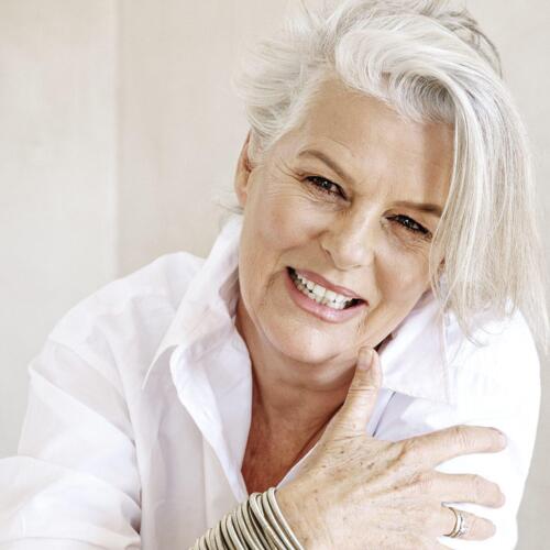 Smiling older woman with white hair wearing a white shirt and multiple silver bangles, conveying warmth and style