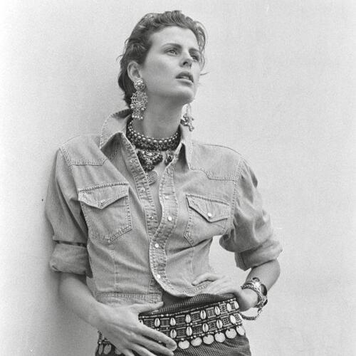 Stylish woman wearing layered necklaces, statement earrings, and a denim button-up shirt, evoking a chic, relaxed vibe.